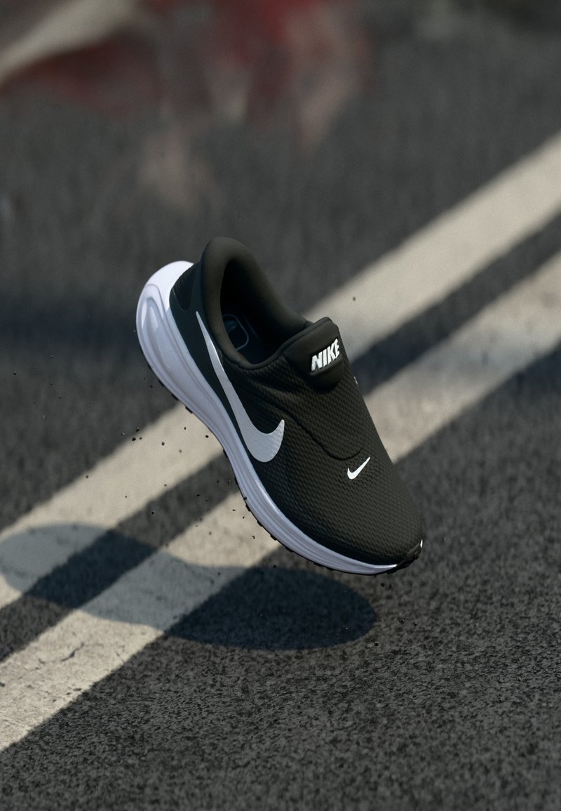 Μαύρο αθλητικό παπούτσι τρεξίματος Nike, χωρίς κορδόνια, με λευκό λογότυπο swoosh και λευκή σόλα, αιωρούμενο πάνω από δρόμο με άσφαλτο και λευκές γραμμές.