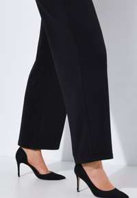 Zwarte wijde broek van rekbaar materiaal, met een gladde textuur en strakke lijnen, gecombineerd met zwarte puntige hakken.