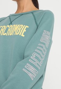 Turkusowy sweatshirt z prążkowanym dekoltem, żółty napis "Abercrombie" z przodu i pionowy biały napis "New York" na lewym rękawie. Miękka mieszanka bawełny.