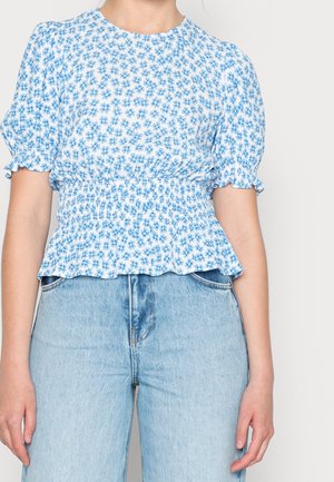 Vrouw draagt een witte blouse met blauw bloemmotief, opgepofte mouwen en een geplooide taille, gecombineerd met lichtblauwe, hooggesneden jeans.