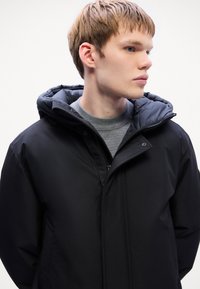 Veste noire imperméable avec un col montant et une capuche amovible, dotée de fermetures éclair et d'une texture lisse et élégante.