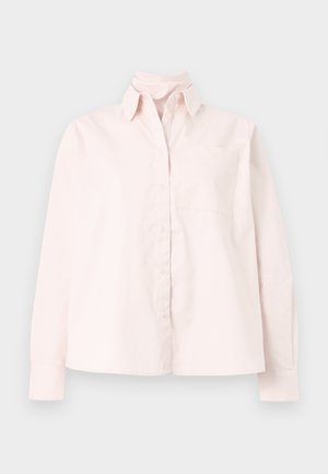 Žena oblečená v bledoružovej košeli na gombíky, modrých širokých džínsach s malými bielymi diamantovými vzormi a čiernych otvorených lodičkách s priesvitnými blokovými podpätkami.