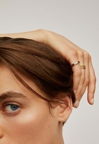 Persona con cabello castaño y ojos azules vista de perfil, con la mano descansando en la cabeza, usando dos anillos en el dedo medio.