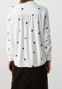 Blusa bianca con piccoli ricami floreali blu, tessuto leggero, maniche lunghe con polsini abbottonati e dettaglio arricciato sul retro.