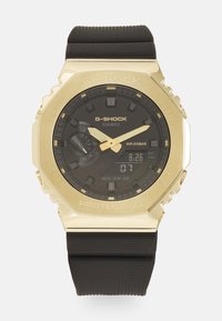 Montre G-Shock avec un cadran noir, boîtier doré et bracelet en caoutchouc noir. Les caractéristiques incluent un écran numérique et des accents de surface texturée.