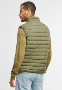 Olivgrüne Pufferweste mit gestepptem Design, hohem Kragen und Reißverschluss, getragen über einem hellbraunen Langarmhemd und blauen Jeans.