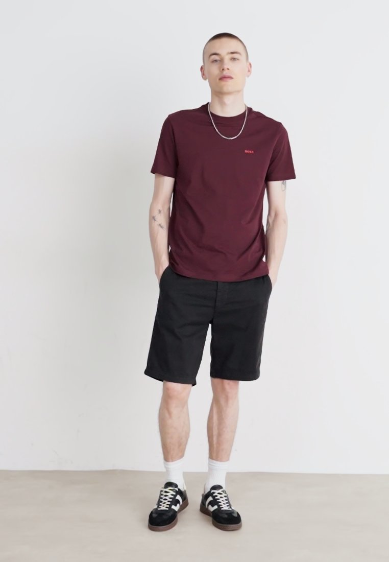 T-shirt en coton bordeaux avec un petit logo, assortie à un short noir. Le modèle porte des baskets noires avec des accents blancs et des chaussettes blanches mi-mollet.