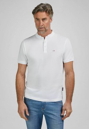Homme d'âge moyen aux cheveux gris portant un t-shirt henley blanc à manches courtes et un jean bleu, debout devant un fond gris uni.