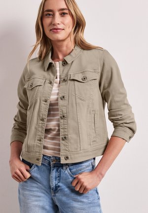 Frau trägt eine beige, geknöpfte Jacke über einem gestreiften Hemd und hellblaue Jeans, steht mit den Händen in den Taschen vor einem einfarbigen Hintergrund.