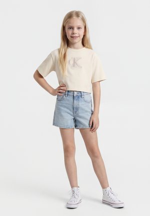 Joven chica con pelo largo y rubio que lleva una camiseta recortada beige, pantalones cortos vaqueros azul claro y zapatillas blancas de caña alta, de pie con una mano en la cadera.