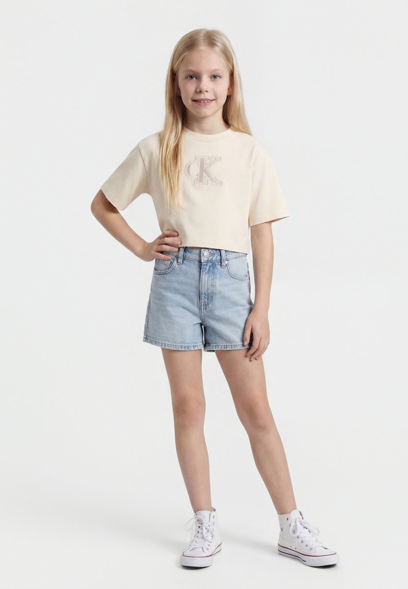 Noor tüdruk pikkade blondide juustega, kandes beeži lühendatud t-särki, helerohelisi teksapükse ja valgeid kõrgetel põlvikutega spordijalatsid, seistes ühe käega puusal.