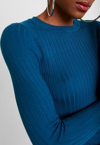 Haut côtelé bleu sarcelle à manches longues avec une silhouette ajustée, présentant une texture subtile et un plissage aux épaules.