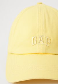 Gul baseballcap med brodert "GAP"-logo. Laget av teksturert stoff med buet skygge og toppknappdetalj.