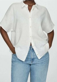 Biała koszula z krótkim rękawem wykonana z lekkiego materiału, z zapięciem na guziki z przodu, luźnym krojem i widocznym przeszyciem. Połączona z jasnoniebieskimi dżinsami.