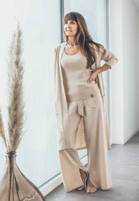 Beige gestricktes Tanktop, langer Cardigan und weitere Hosen mit Bindeband. Glatte Textur und helle Farben vor einem minimalistischen Hintergrund.