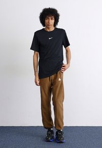 Crna majica kratkih rukava marke Nike u kombinaciji sa smešnim sportskim hlačama smeđe boje koje imaju beli logo, upotpunjene crnim patikama s plavim detaljima.