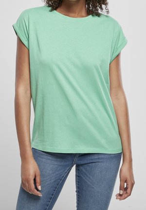 Kvinde iført en kortærmet mintgrøn T-shirt og blå jeans, stående med afslappede arme mod en ensartet lys baggrund.