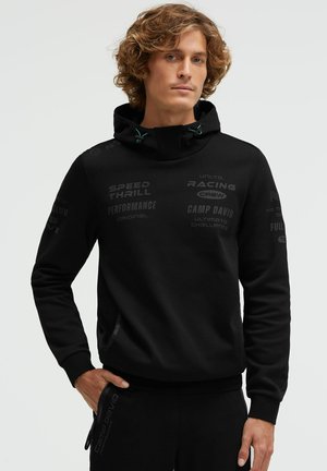 Man with curly hair wearing black hoodie and pants with subtle racing text print, standing with one hand in pocket against plain background.