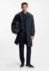 Abrigo impermeable azul marino con capucha, bolso negro texturizado, camiseta azul marino, pantalones oscuros y zapatillas deportivas oscuras. Presenta un diseño ajustado y herrajes sencillos.