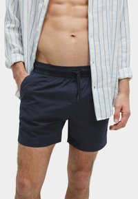 Bañadores azul marino con cintura elástica y cordón, con bolsillos laterales, combinados con una camisa blanca de rayas y botones.