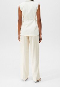 Ärmelloses cremefarbenes Oberteil mit tailliertem Gürtel, kombiniert mit weiten cremefarbenen Hosen. Das Material wirkt weich und hat eine glatte Textur.
