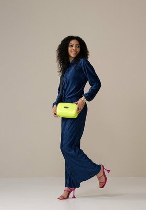 Marineblauwe geplooide jurk met een ceintuur in de taille, gecombineerd met een limoen groene crossbody tas en roze bandjeshakken. Model staat op een lichte ondergrond.