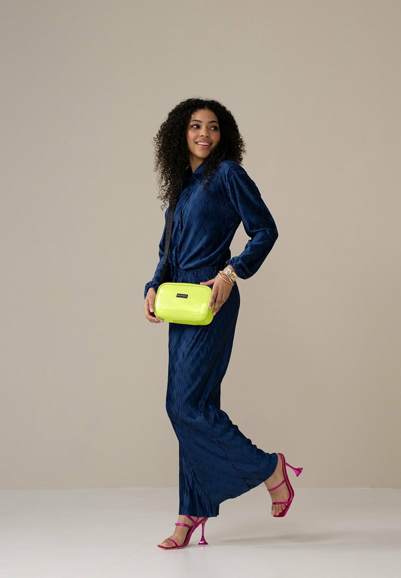 Marineblaues, plissiertes Kleid mit einem taillierten Gürtel, kombiniert mit einer limegrünen Umhängetasche und pinken Riemchenabsätzen. Das Model steht auf einer hellen Fläche.