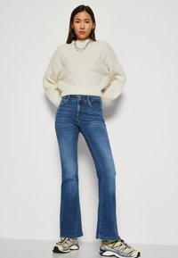 Hvid chunky striktrøje, højtaljede blå flared jeans, tekstureret stof, subtile syninger, parret med beige og sorte sporty sko.