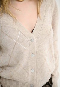 Cardigan beige con scollo a V, caratterizzato da un motivo a diamante cucito con filo metallico. Ha cinque bottoni e orli a coste.