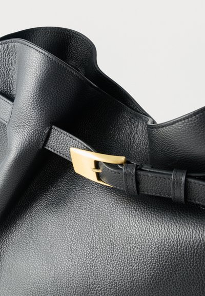 Μαύρη τσάντα από δέρμα με υφή, με χρυσή ορθογώνια αγκράφα σε ρυθμιζόμενο λουρί, σε φωτεινό φόντο.