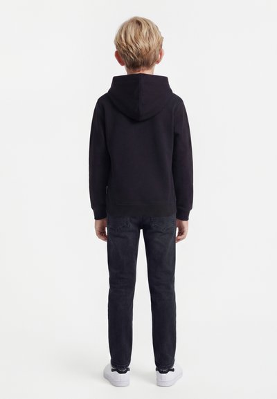 Αγόρι με ξανθά μαλλιά στέκεται με την πλάτη προς τα έξω, φοράει μαύρο φούτερ με κουκούλα, σκούρα τζιν και λευκά αθλητικά παπούτσια σε λευκό φόντο.