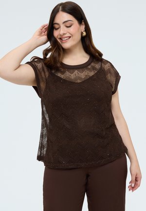 Femme souriante aux cheveux châtain mi-longs porte des boucles d'oreilles créoles dorées, un haut en dentelle marron foncé à manches courtes et un pantalon assorti sur fond clair.