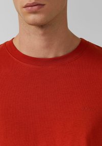 Rotes Baumwoll-T-Shirt mit Rundhalsausschnitt und kurzen Ärmeln. Verfügt über ein kleines gesticktes Logo in der unteren linken Ecke. Glatte Textur.