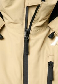 Beige Freizeitjacke mit schwarzen wasserdichten Reißverschlüssen, Reißverschlusszippern und verstellbaren Kordelzügen an den Front- und Brusttaschen.
