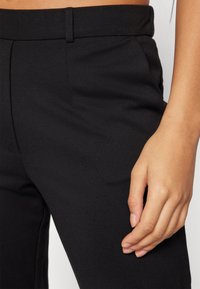 Svarta skräddarsydda byxor med en jämn textur, med en platt framdel, sidofickor och en åtsittande midja; närbild av tyget och handen.