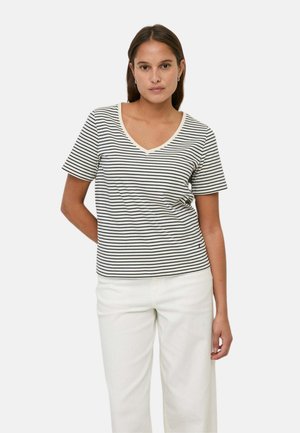 Mujer con camiseta blanca de manga corta y escote en V con rayas horizontales negras y pantalones blancos, de pie frente a un fondo liso.