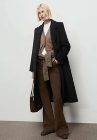 Abrigo largo negro sobre un cárdigan marrón y una camisa blanca, combinado con pantalones acampanados marrones. Los accesorios incluyen un bolso a juego y un cinturón texturizado.