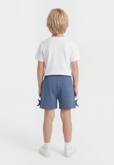Enfant aux cheveux blonds vu de dos, portant un t-shirt blanc, un short bleu, des chaussettes blanches et des baskets blanches sur un fond uni.