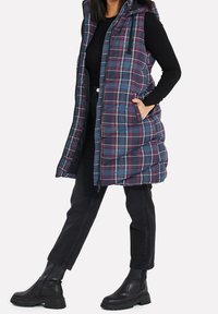 Prošívaná vesta s kostkovaným vzorem v námořnické modři, burgundské a tyrkysové barvě, s kapucí, předním zipem a kapsami. Nositá přes černý dlouhý rukáv a černé džíny.