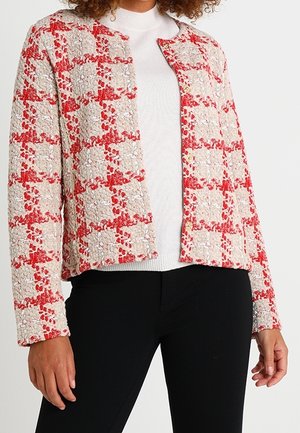 Kvinde iført en rød og beige ternet tekstureret jakke over en hvid sweater og sorte bukser, stående med hænderne afslappet ned langs siderne.