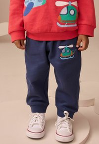 Bambino che indossa pantaloni blu navy e una felpa rossa con toppe di elicotteri verdi e blu, in piedi su piattaforme circolari beige con scarpe da ginnastica bianche.