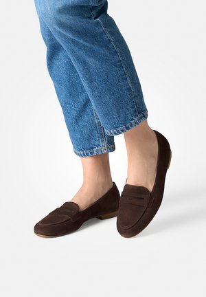 Füße in dunkelbraunen Wildleder-Loafers, kombiniert mit blauen, gekürzten Jeans auf weißem Hintergrund.