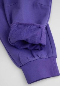 Fioletowe dresy z gładką teksturą, elastycznymi mankietami i bocznymi kieszeniami. Materiał wygląda na miękki i wygodny.