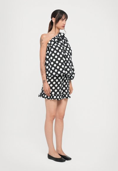 Schwarzes und weißes Polka-Dot-Zweiteiler-Outfit mit einem lockeren Oberteil und einem figurbetonten Rock. Das Oberteil hat ein geknotetes Detail; der Rock verfügt über einen gerafften Saum.