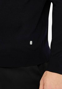 Maglione lavorato a maglia blu navy con orlo e polsini a coste, con una piccola accentuazione del logo argento sul lato sinistro inferiore.