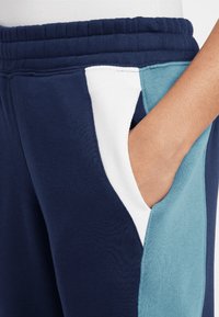 Námornícky modré tepláky s bielou a tyrkysovou bočnou kapsou. Materiál je jemný; dizajn obsahuje elastický pás a zúženú nohu.