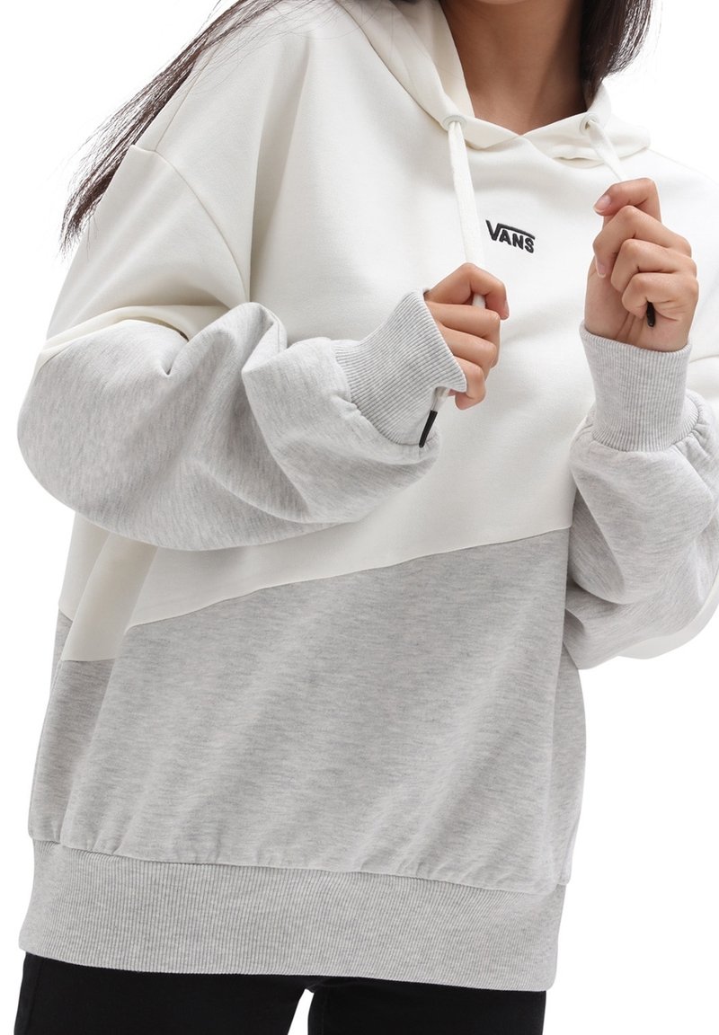 Femme portant un sweat à capuche Vans blanc et gris clair, ajustant les cordons près de la poitrine avec les deux mains.