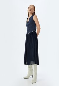 Falda plisada azul marino combinada con un chaleco ajustado azul marino con un sutil patrón de rayas y borde blanco; botas altas blancas hasta la rodilla.