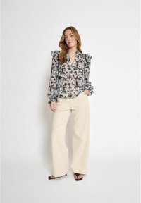 Chemisier transparent avec un motif abstrait gris et beige, épaules volantes et décolleté en V. Associé à un pantalon large crème et des sandales noires.