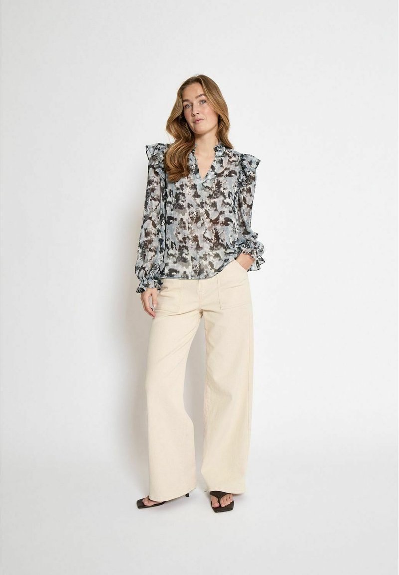 Chemisier transparent avec un motif abstrait gris et beige, épaules volantes et décolleté en V. Associé à un pantalon large crème et des sandales noires.